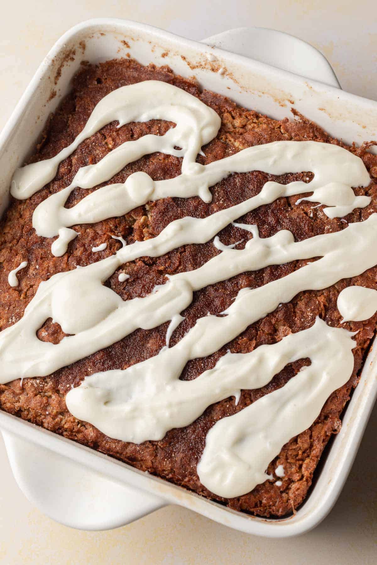 A baked cinnamon oatmeal square in a white dish, topped with drizzled white icing.