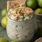 A jar of key lime pie overnight oats topped with crushed graham crackers and lime zest, surrounded by limes.