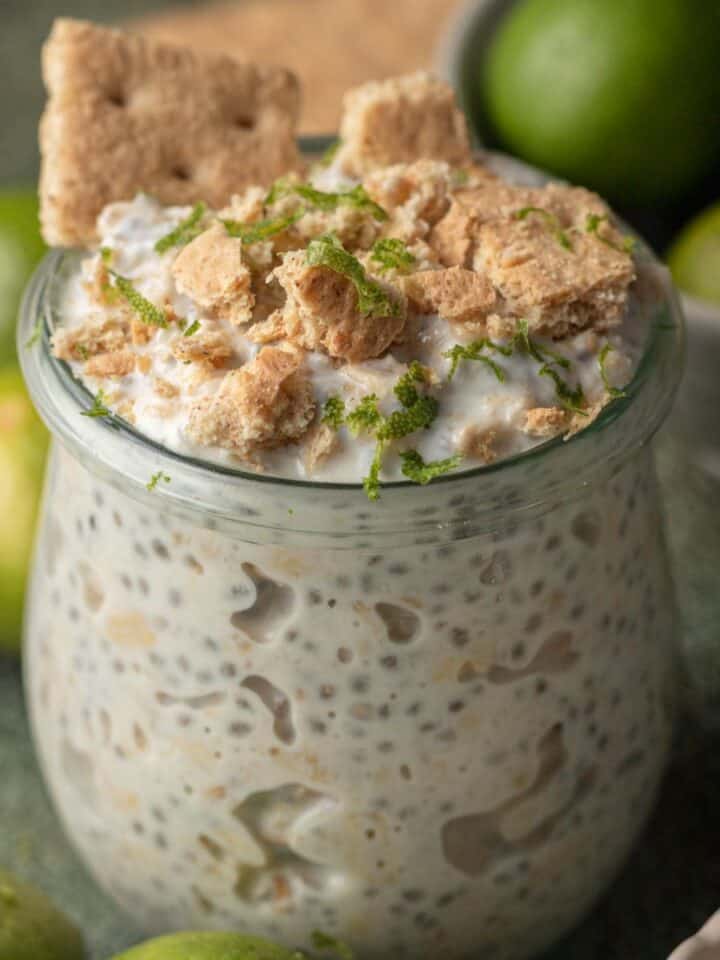 A jar of key lime pie overnight oats topped with crushed graham crackers and lime zest, surrounded by limes.