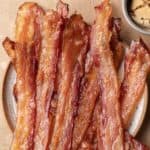 Crispy cooked bacon strips on a plate with a small bowl of brown sugar nearby.