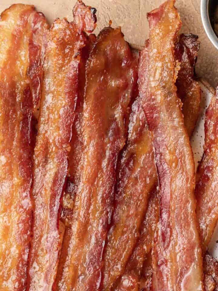 Crispy cooked bacon strips on a plate with a small bowl of brown sugar nearby.