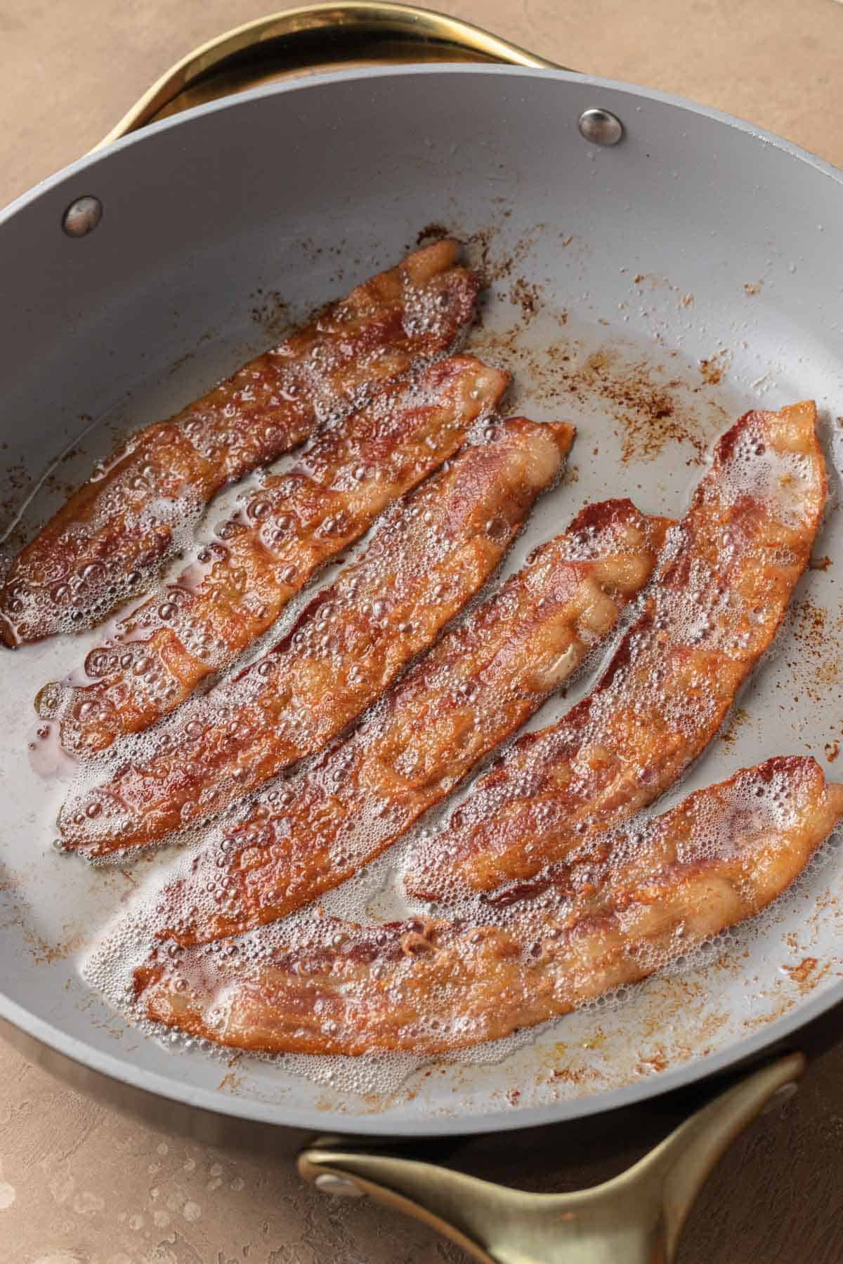 Four strips of bacon sizzling and browning in a frying pan with bubbling grease.