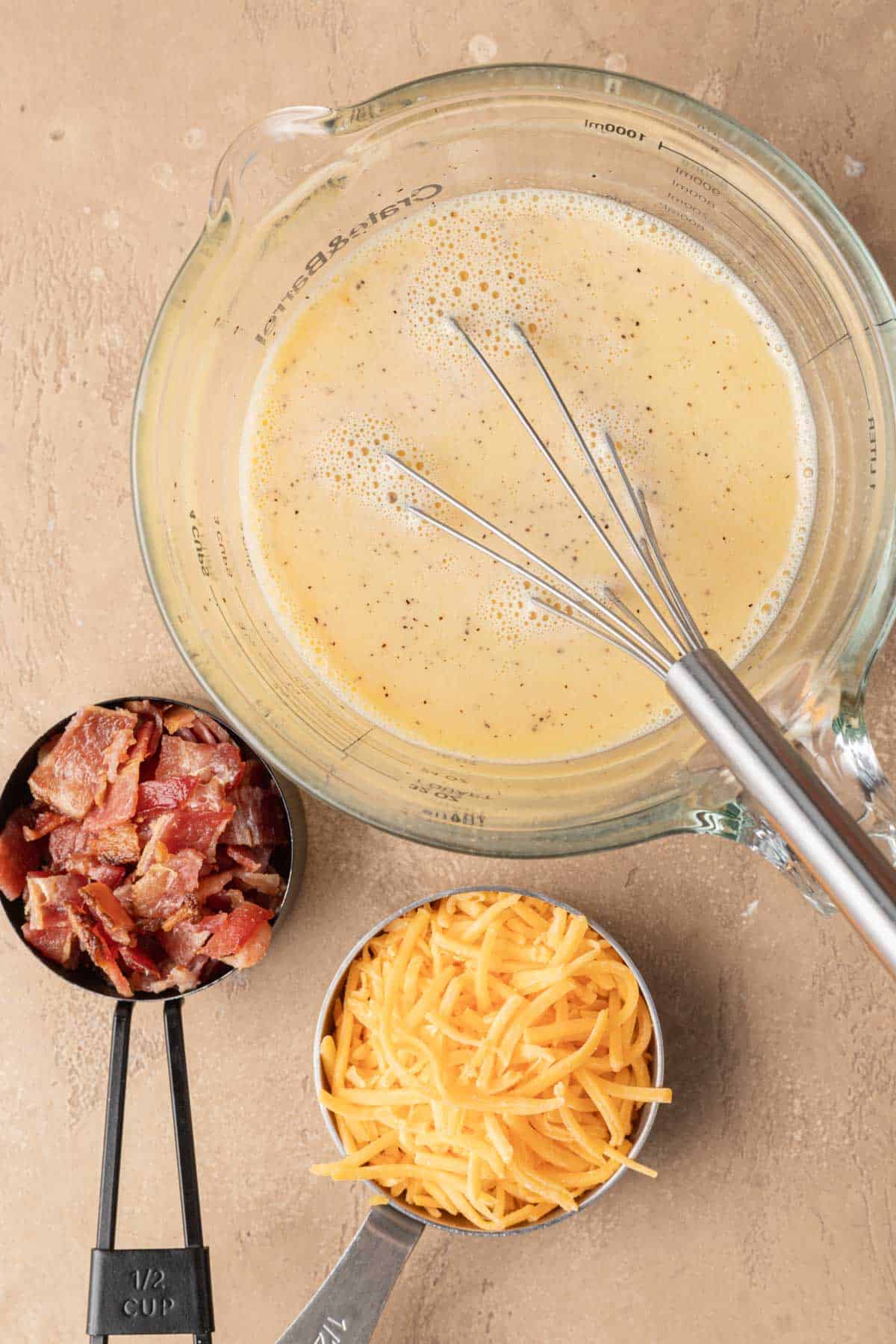 A bowl of whisked eggs with a metal whisk, next to cups of cooked bacon pieces and shredded cheddar cheese.