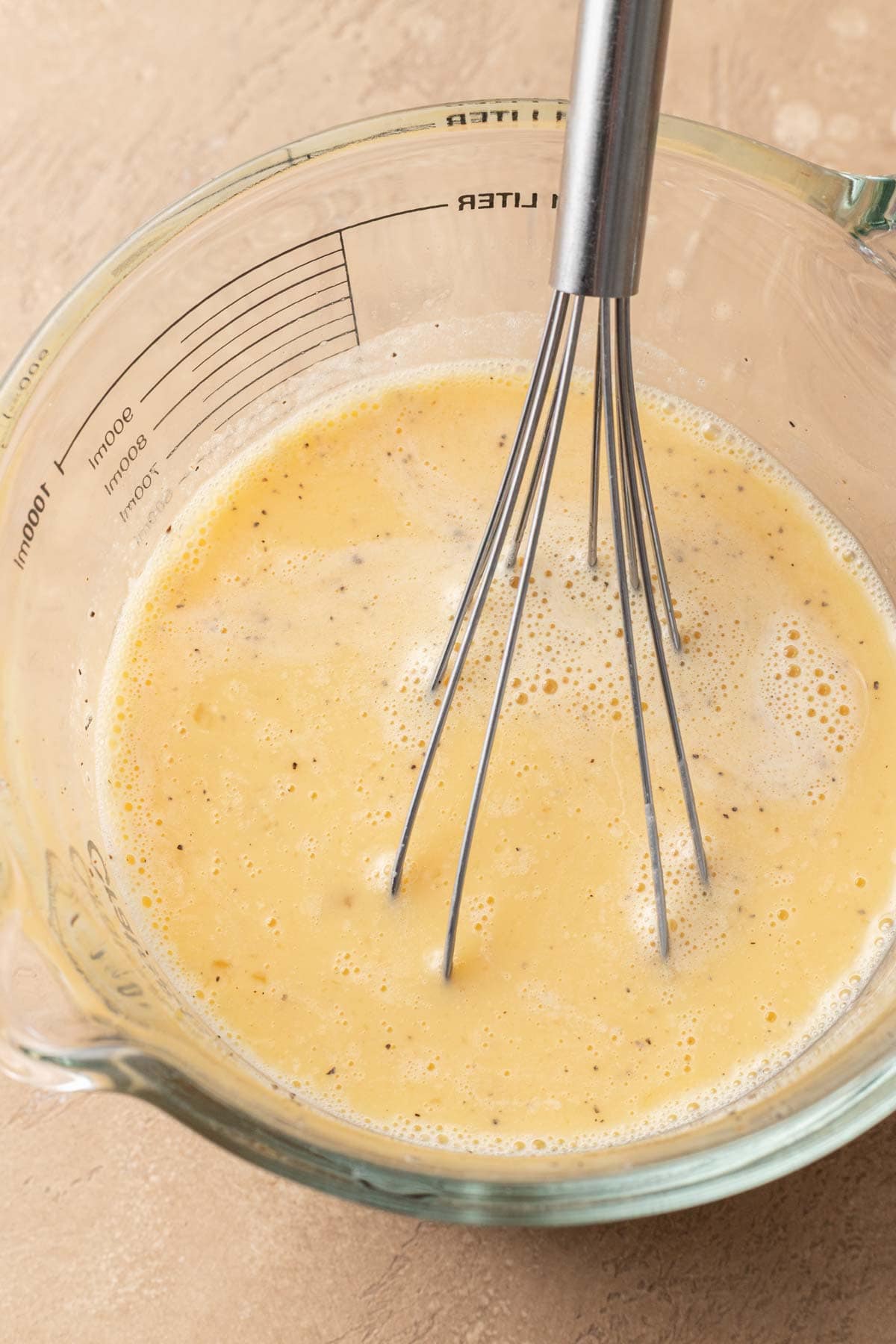 A metal whisk mixing beaten eggs in a clear glass measuring cup on a beige surface.
