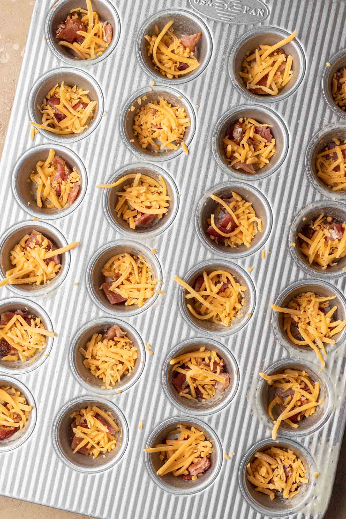 A muffin tin filled with bacon pieces and shredded cheddar cheese in each cup, ready to be baked.