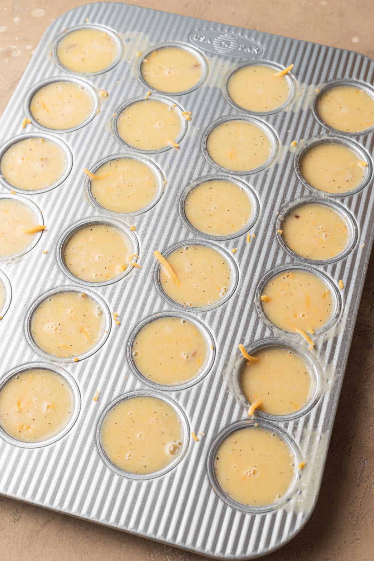 A muffin tin filled with uncooked egg mixture, ready to be baked, on a beige surface.