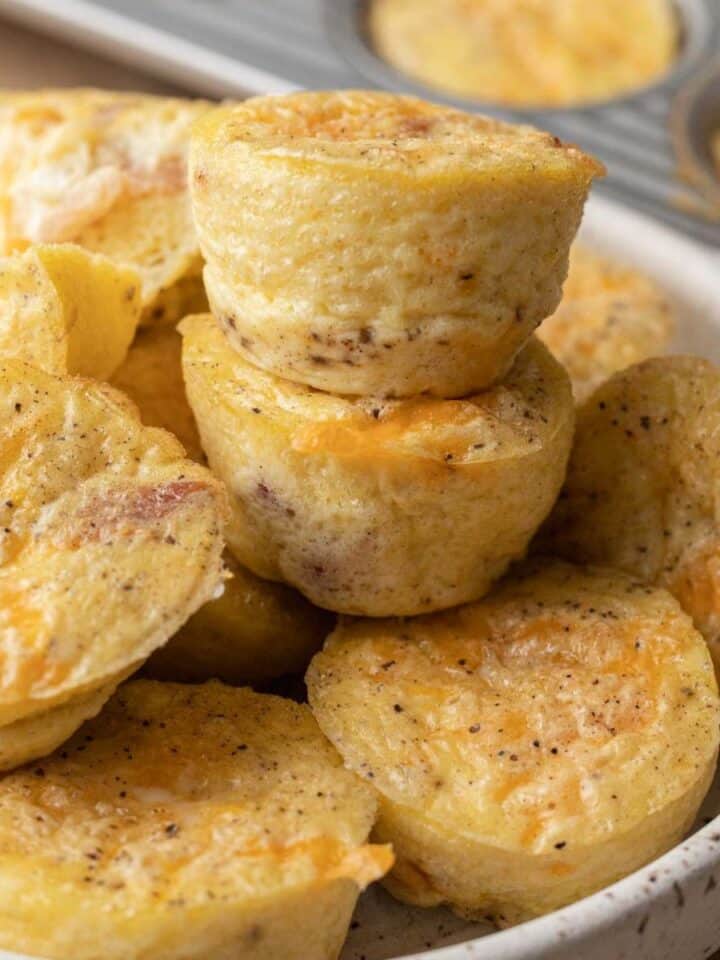 Mini egg muffins with cheese and bacon stacked in a bowl, with a muffin tin in the background.