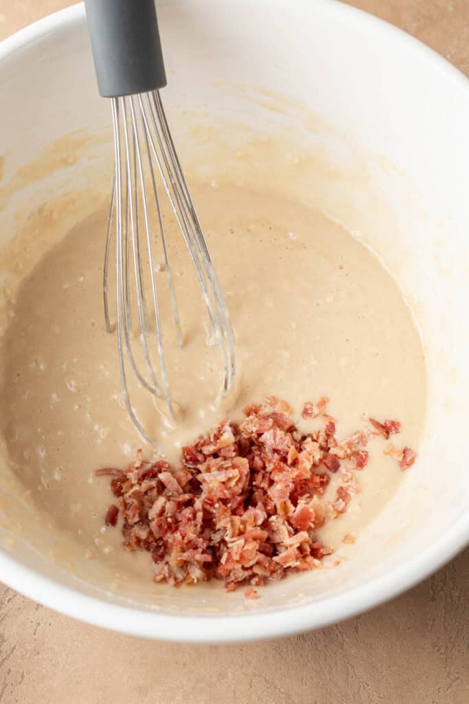 A whisk in pancake batter with chopped cooked bacon in a white mixing bowl.