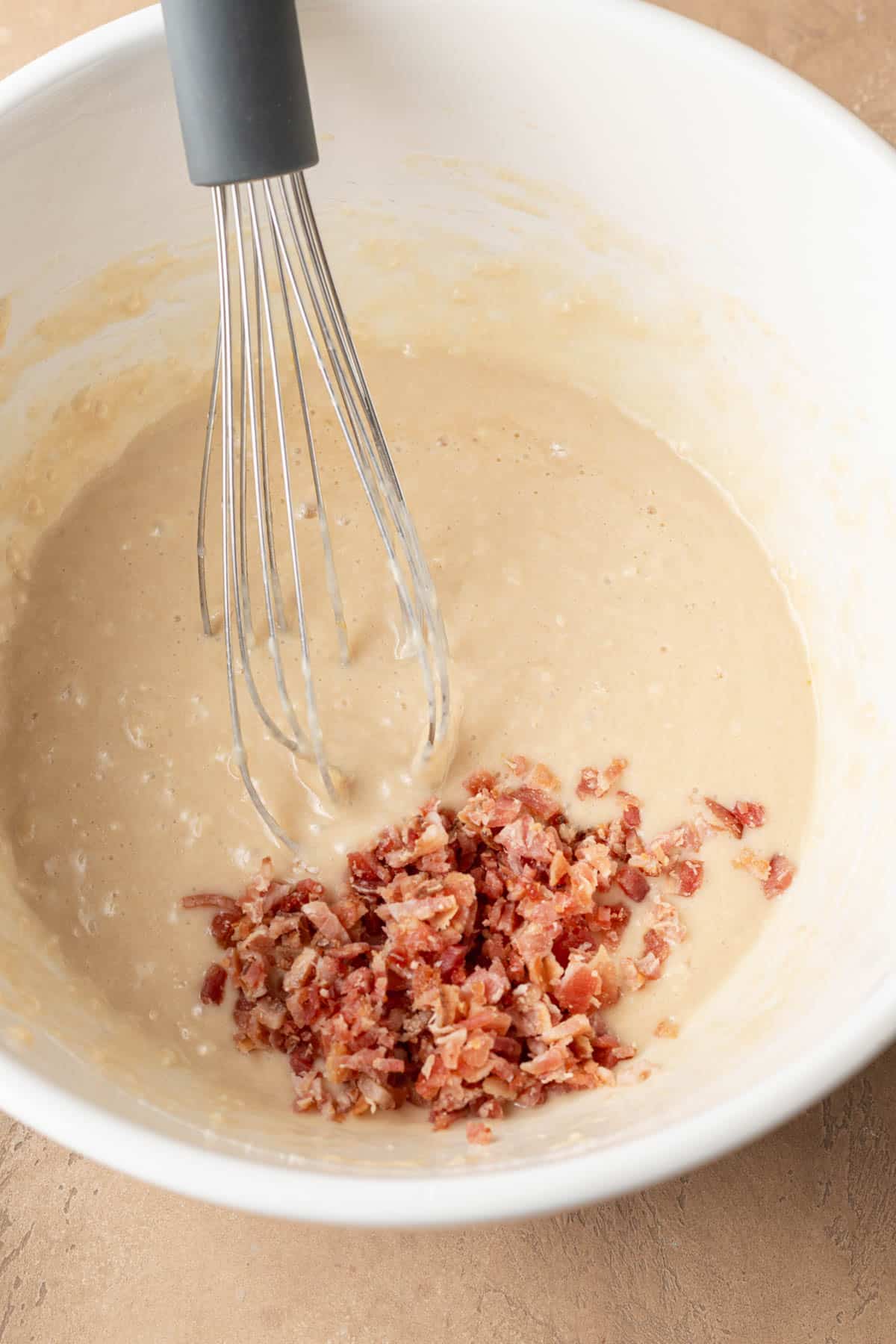 A whisk in pancake batter with chopped cooked bacon in a white mixing bowl.