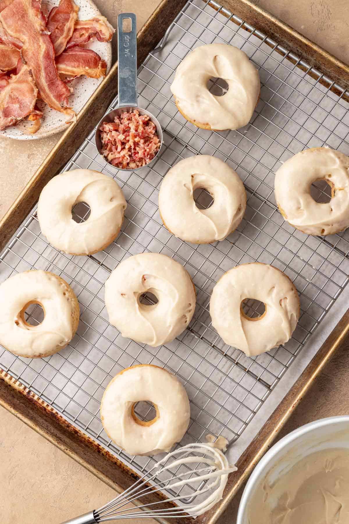Eight glazed donuts on a cooling rack, with bacon bits and cooked bacon slices nearby.