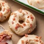 A donut with maple glaze and bacon bits on top, resting on a brown surface next to more donuts and bacon pieces.