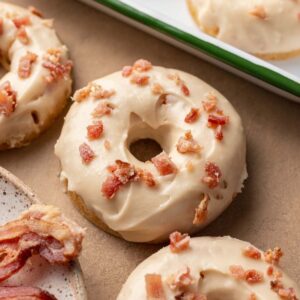 A donut with maple glaze and bacon bits on top, resting on a brown surface next to more donuts and bacon pieces.