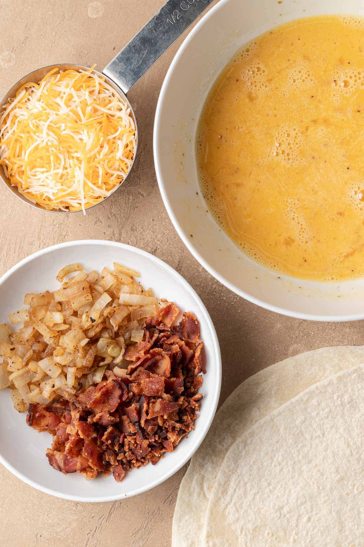 Shredded cheese, beaten eggs, cooked bacon with onions, and flour tortillas arranged on a tan surface.