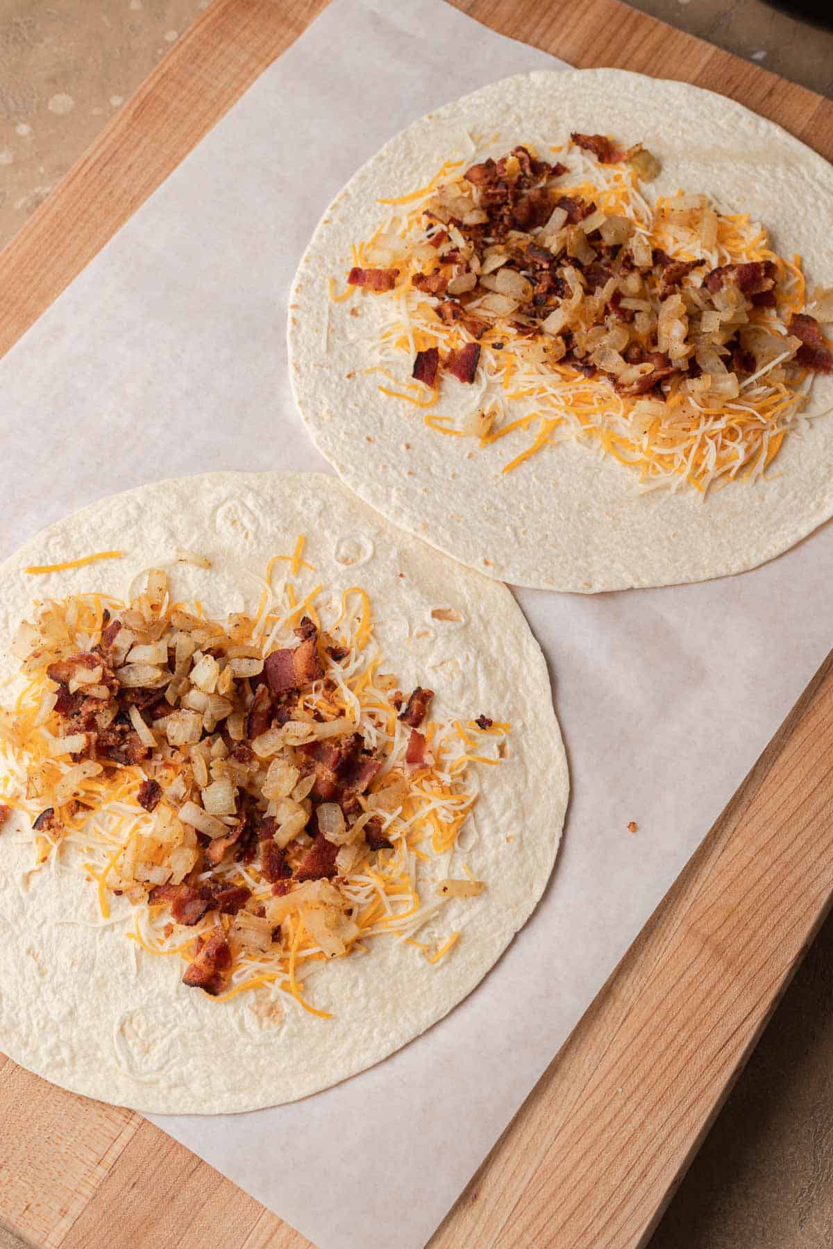Two tortillas on parchment paper topped with shredded cheese, bacon, and diced cooked onions.