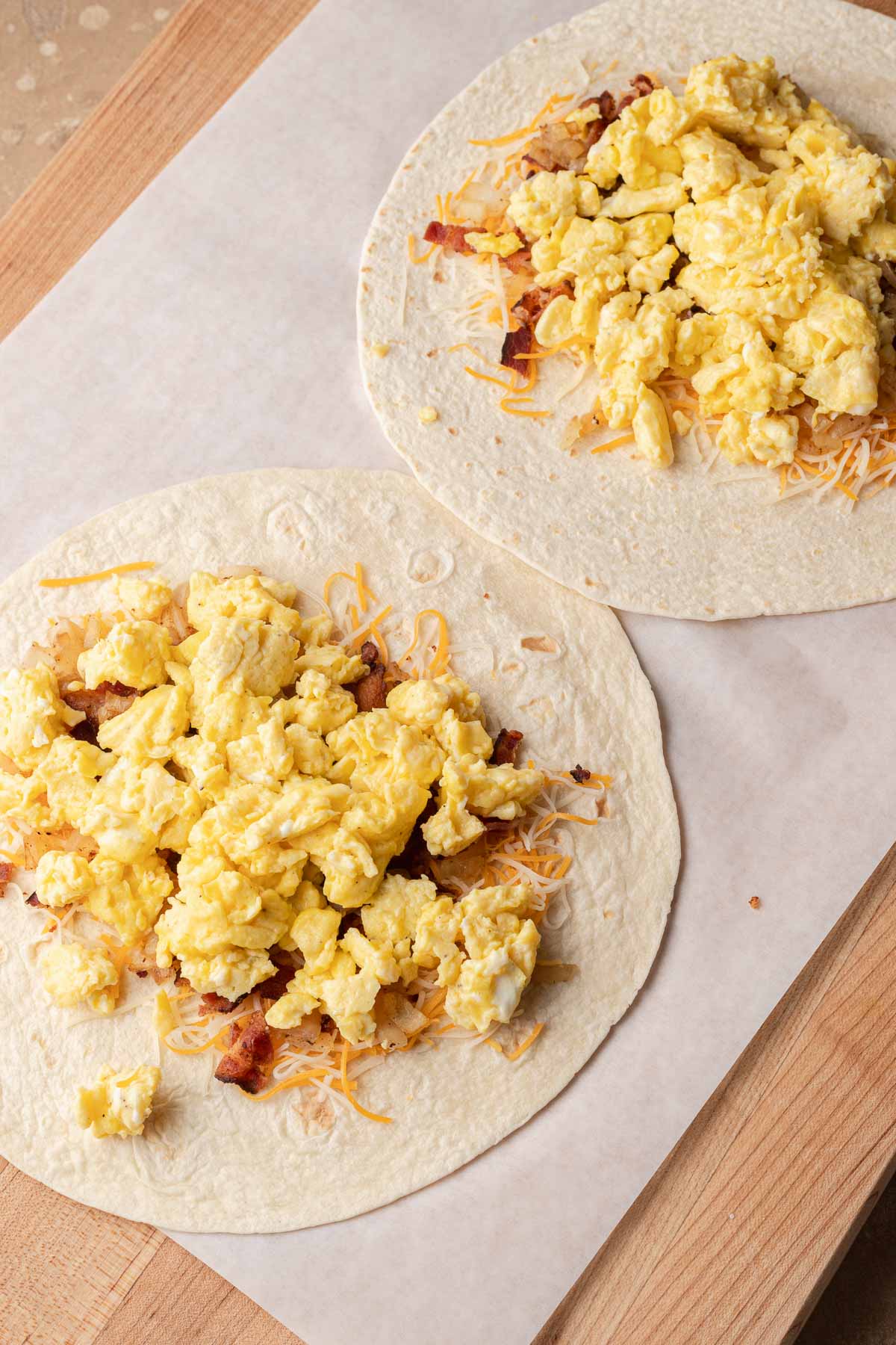 Two flour tortillas topped with scrambled eggs, shredded cheese, and bacon on a wooden surface.