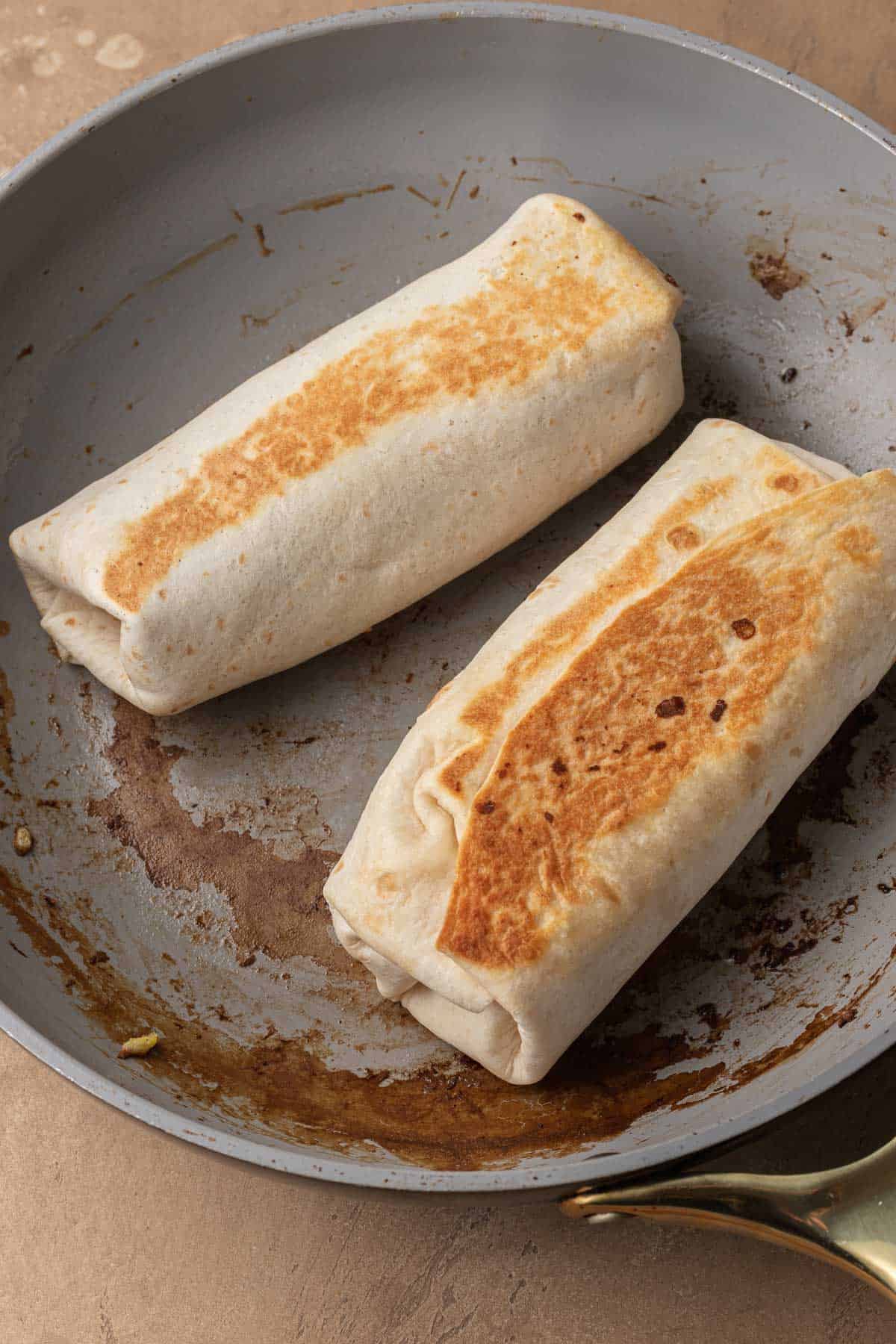 Two golden-brown burritos cooking in a skillet on a stovetop.