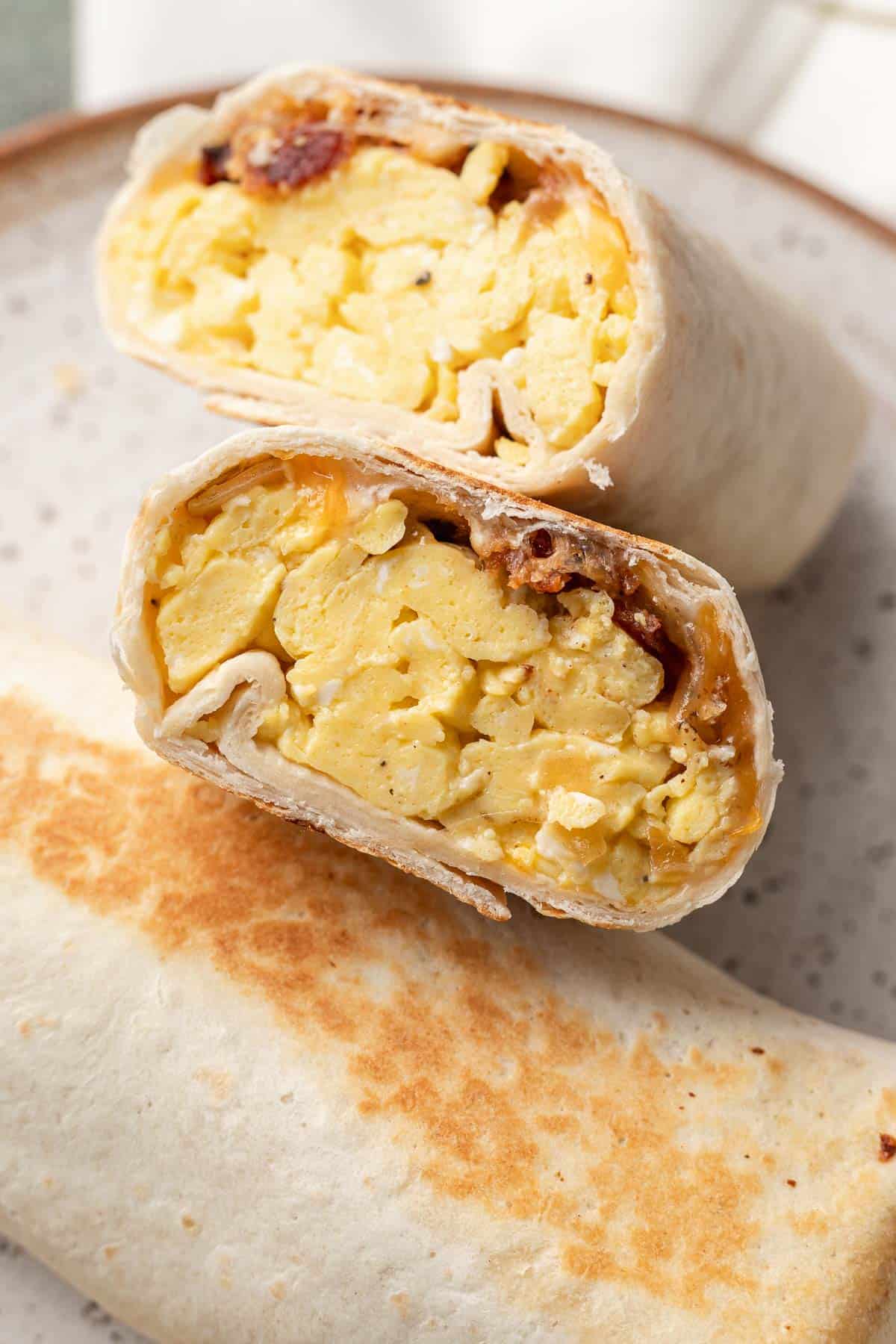 A breakfast burrito cut in half, showing scrambled eggs and bacon inside a tortilla on a plate.