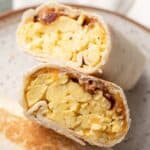 A breakfast wrap cut in half, showing scrambled eggs, bacon, and melted cheese inside a flour tortilla.