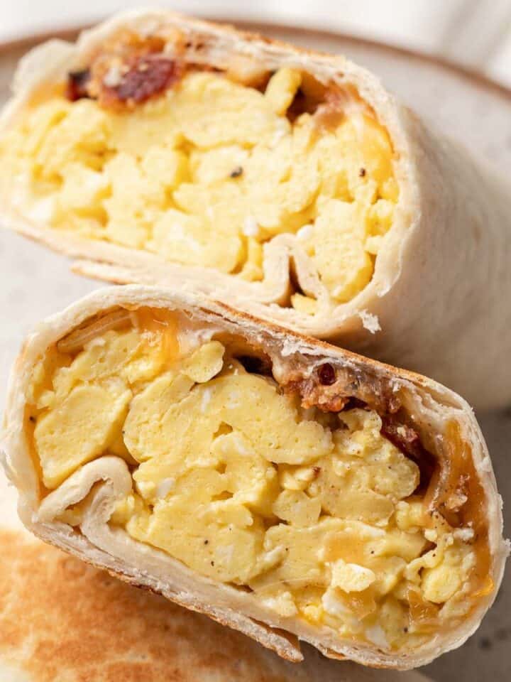 A breakfast wrap cut in half, showing scrambled eggs, bacon, and melted cheese inside a flour tortilla.
