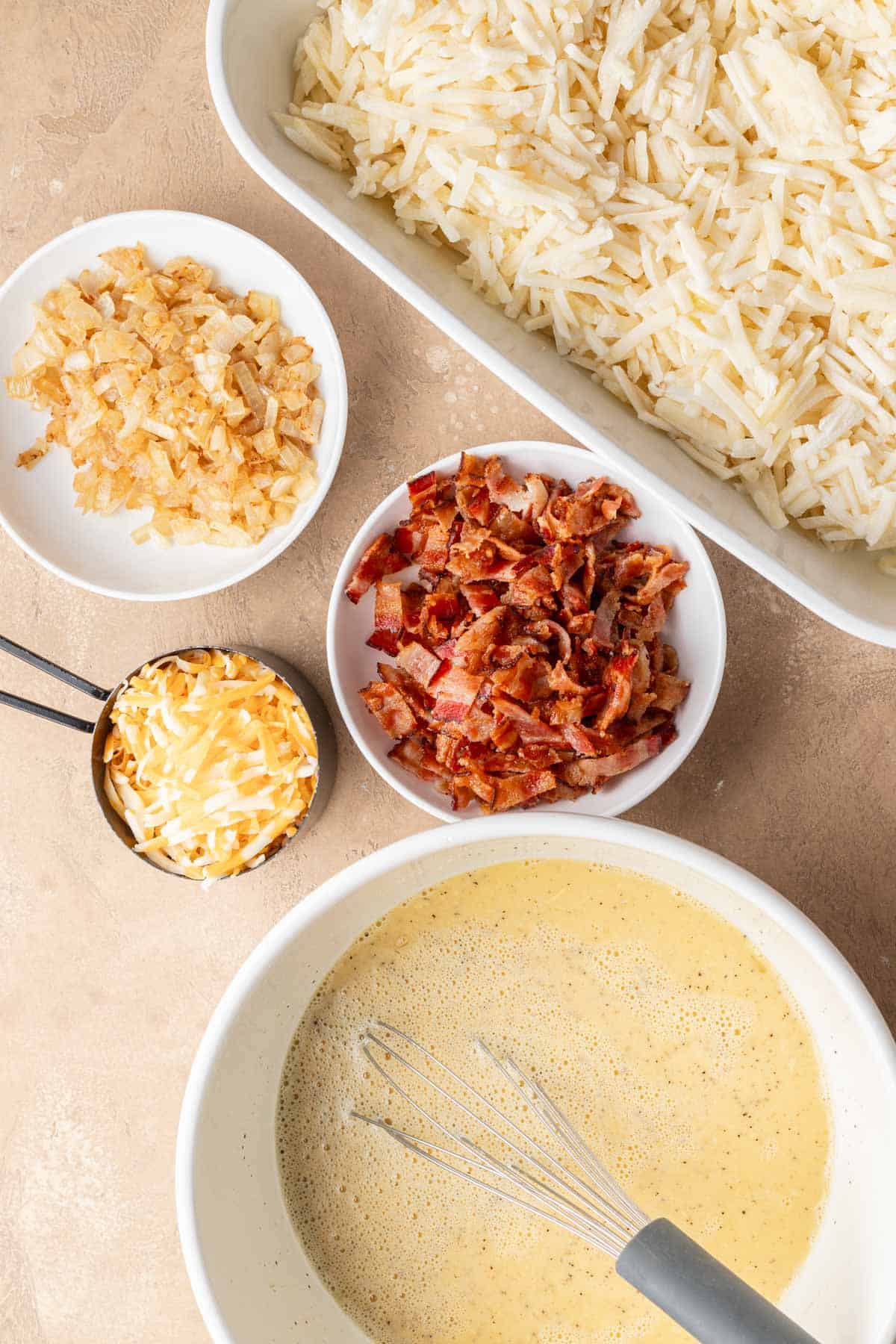 Shredded potatoes, cheese, bacon, onions, and a bowl of whisked eggs are arranged on a countertop.