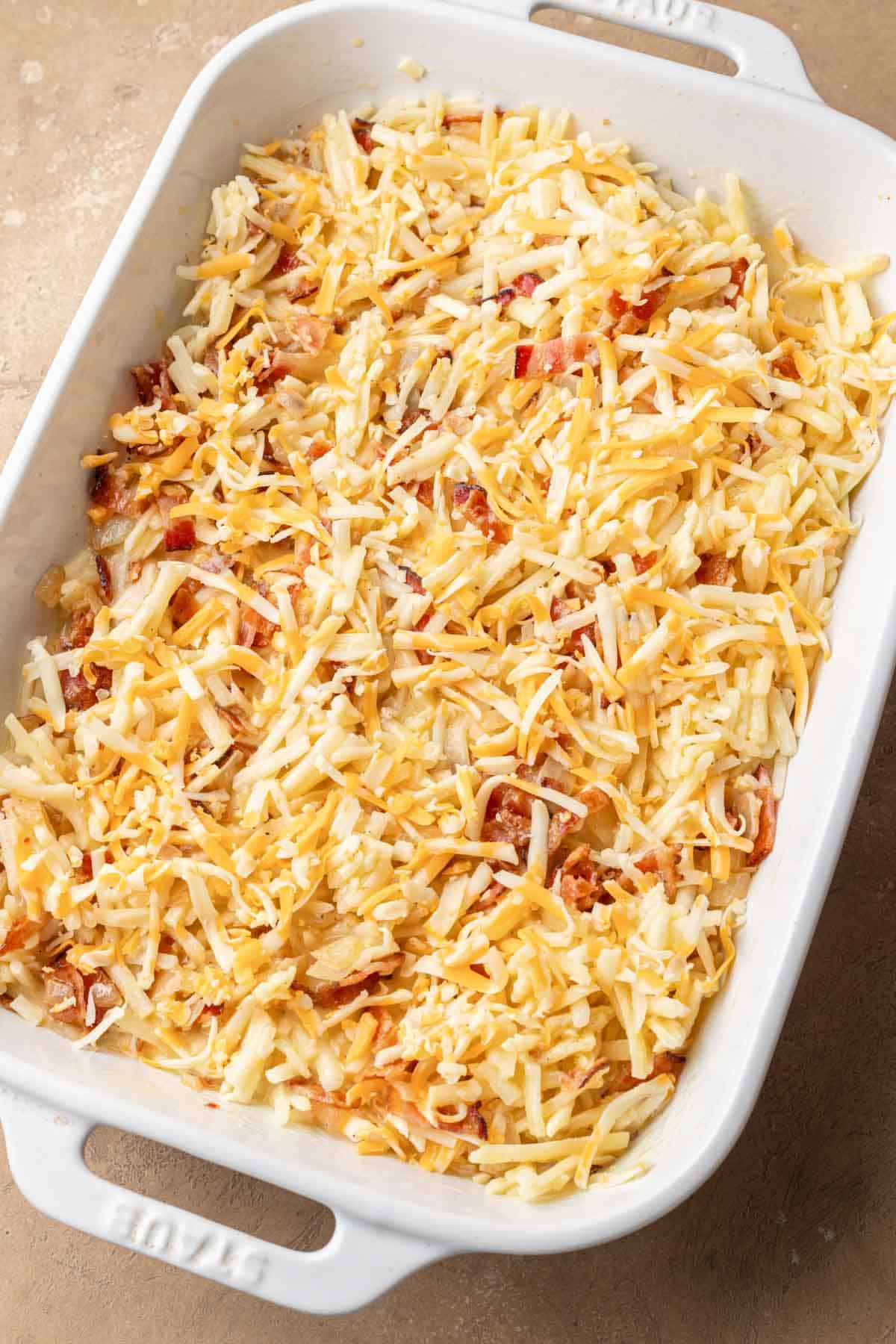 A white baking dish filled with shredded cheese and hash browns, ready to be baked.