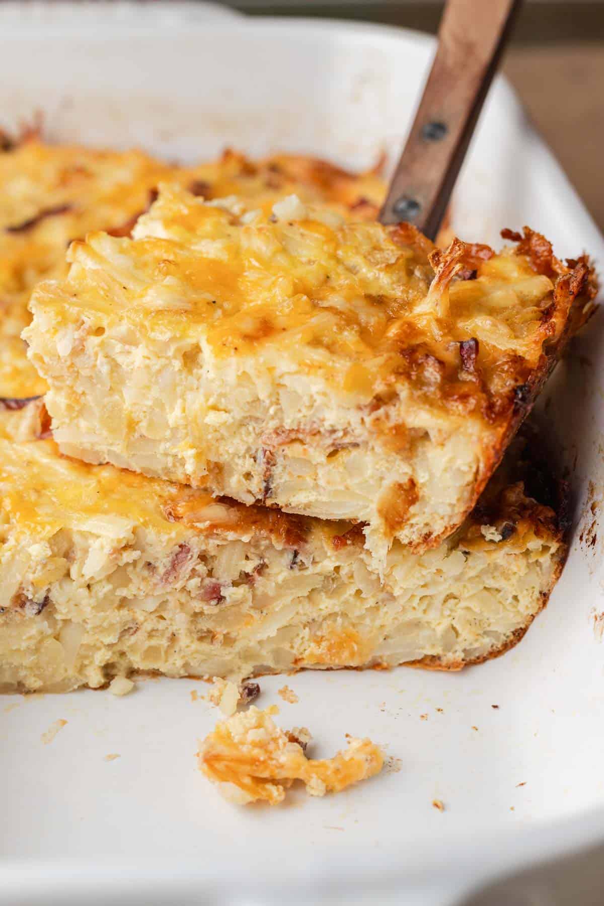 A spatula lifts a cheesy, baked hash brown casserole from a white baking dish.