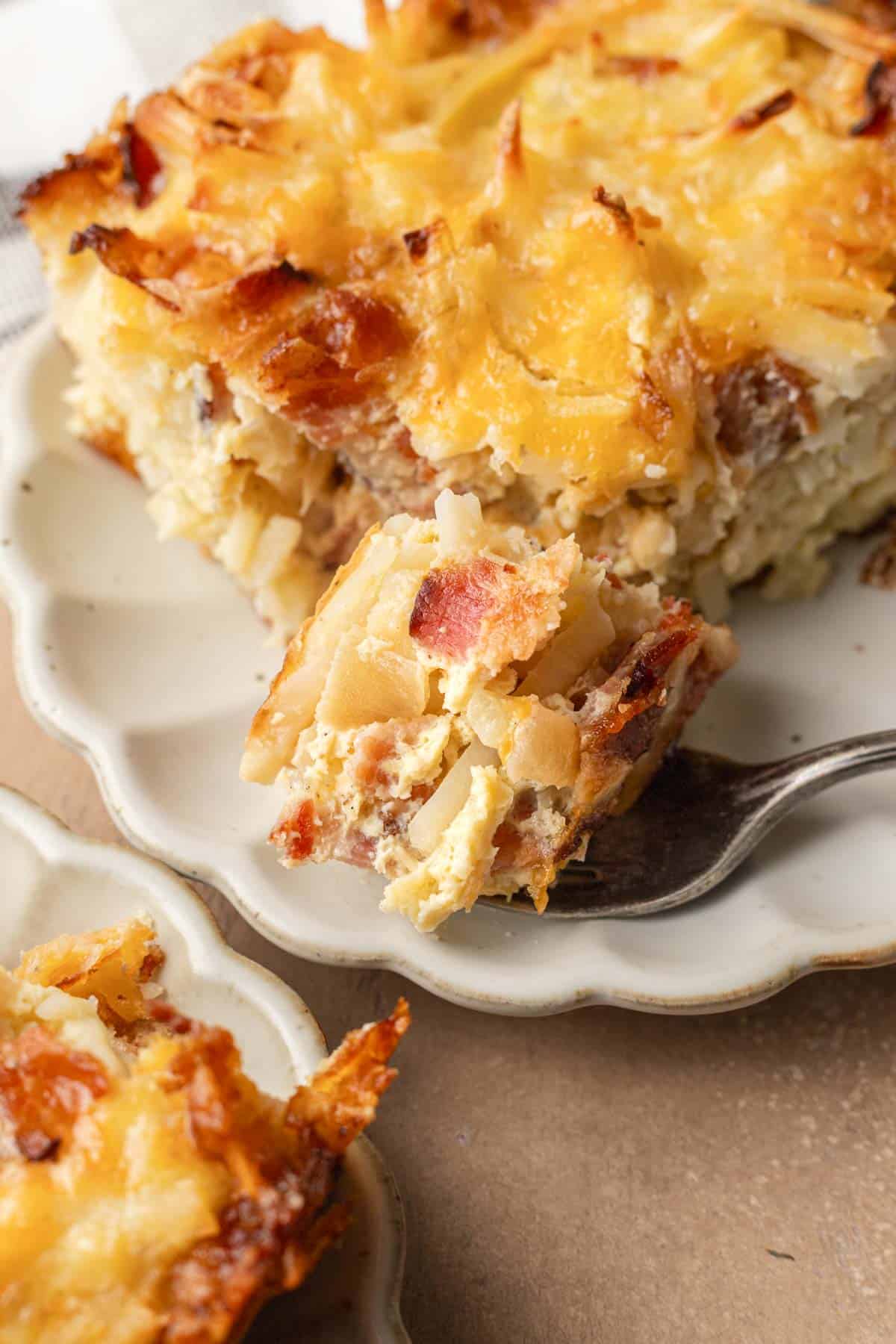 A fork holds a bite of cheesy breakfast casserole with visible bacon, potatoes, and melted cheese on a plate.
