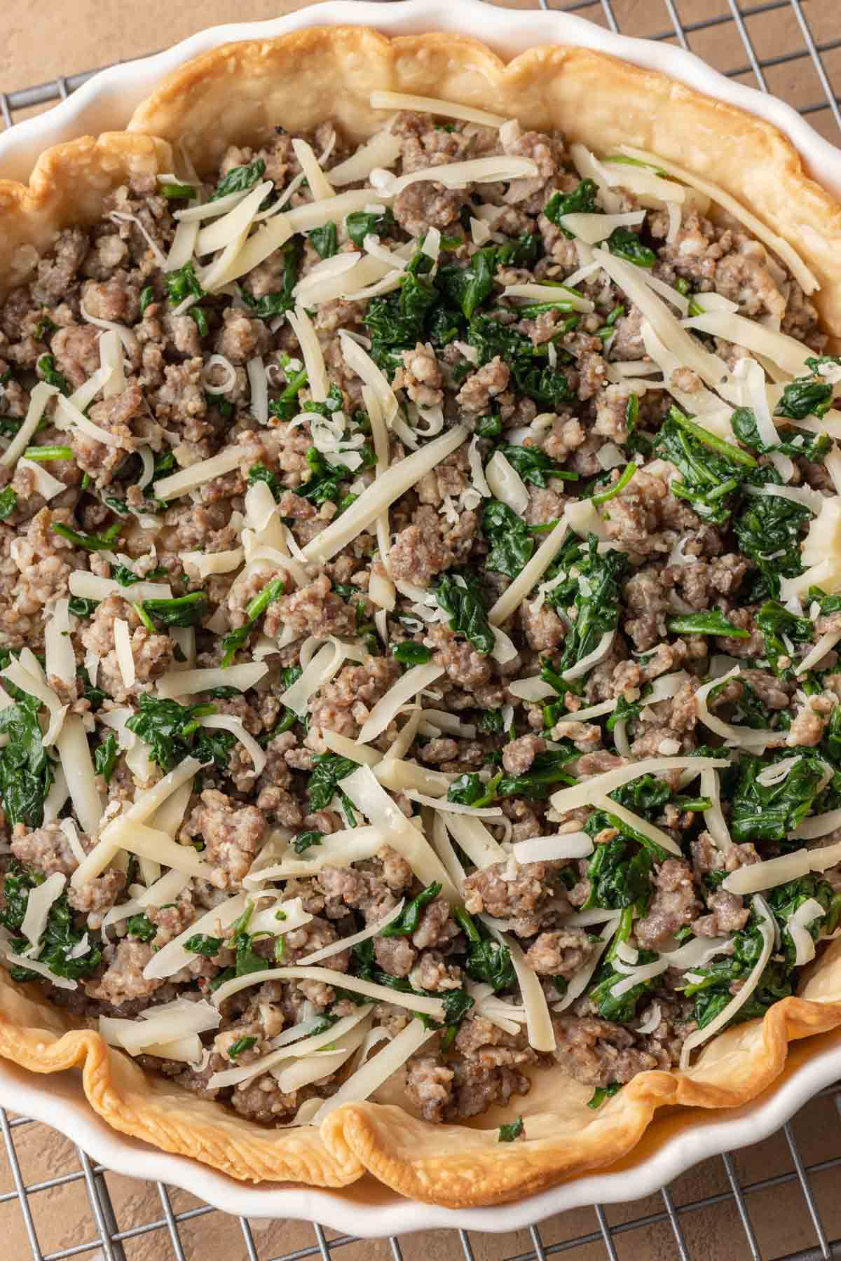 Unbaked pie crust filled with cooked sausage crumbles, shredded cheese, and chopped spinach on a cooling rack.
