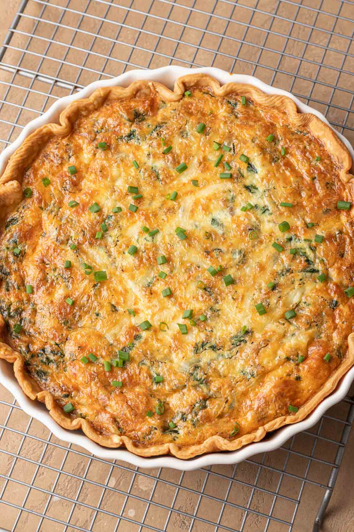 Golden-brown sausage spinach quiche with cheese in a white dish, topped with chopped chives, on a cooling rack.