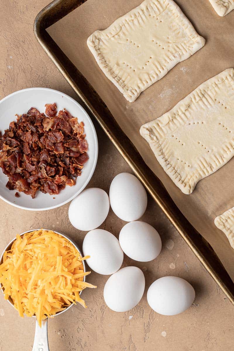 Rectangular pastry dough on a baking sheet, with bacon, shredded cheese, and eggs beside it.