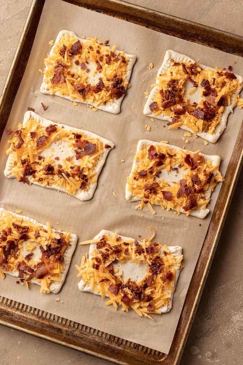 Six unbaked rectangular pastries topped with cheese and bacon on a parchment-lined baking sheet.