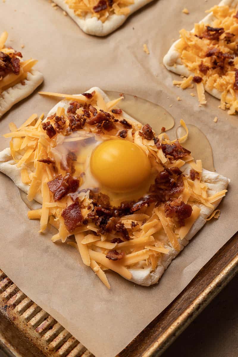 Unbaked breakfast pastry with egg, shredded cheese, and bacon on parchment-lined baking sheet.