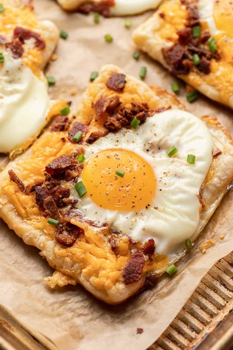 puff pastry breakfast tart topped with melted cheese, a sunny-side-up egg, bacon bits, and chopped chives.