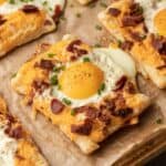 Square breakfast pastries topped with egg, bacon, cheese, and chives on parchment paper.