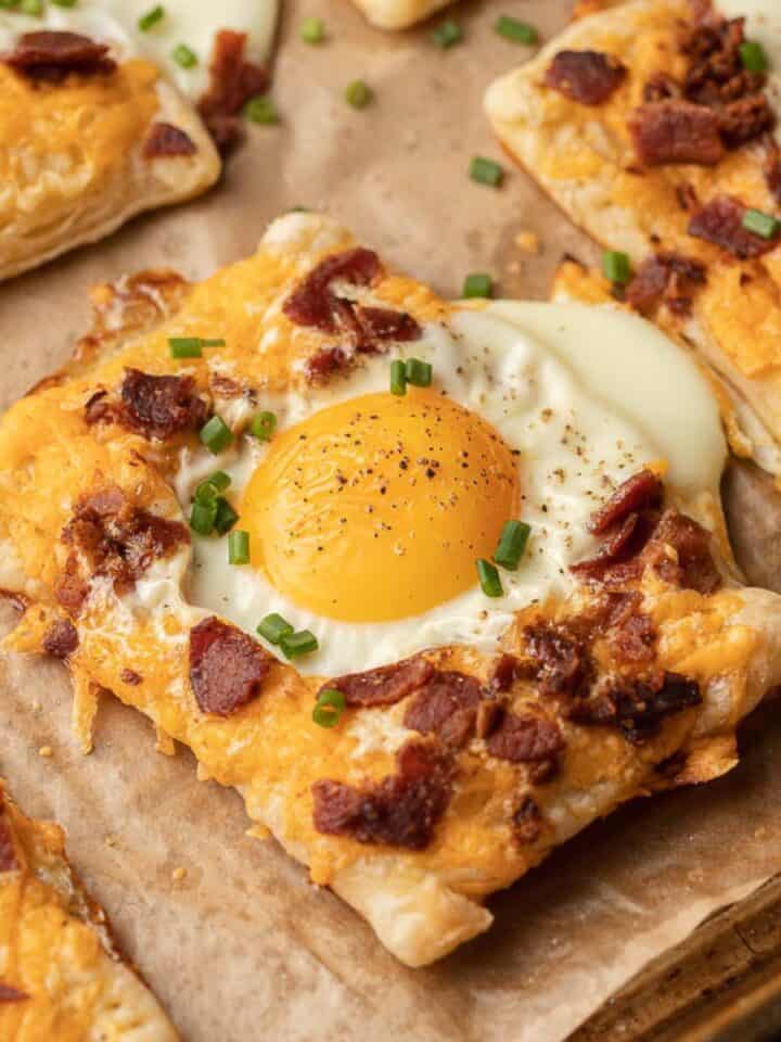 Square breakfast pastries topped with egg, bacon, cheese, and chives on parchment paper.