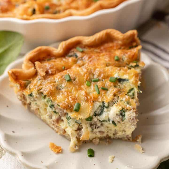 A slice of spinach & sausage quiche with cheese, garnished with chopped chives on a white plate.