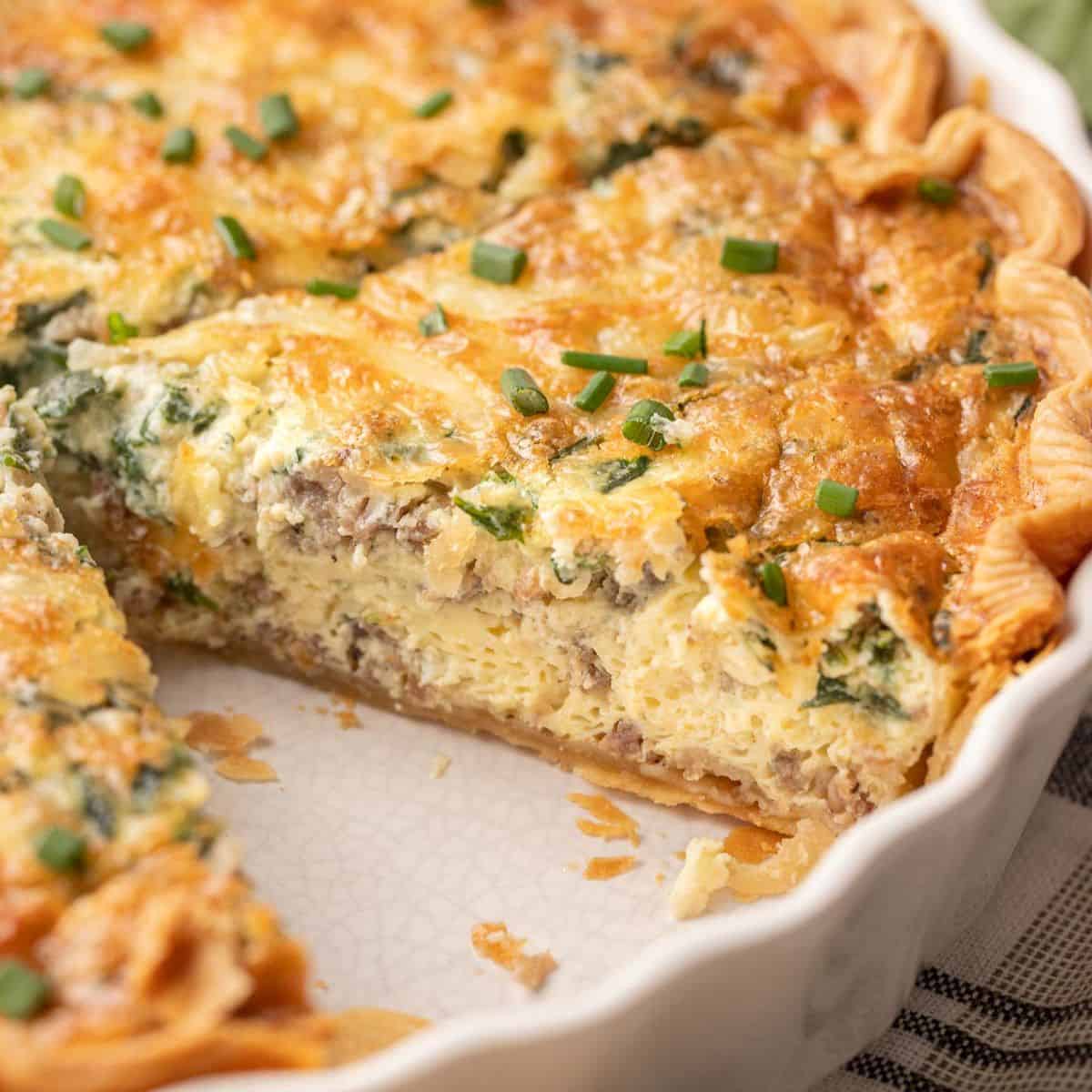 A close-up of a sliced spinach & sausage quiche with a golden crust, creamy filling, and chopped chives on top.