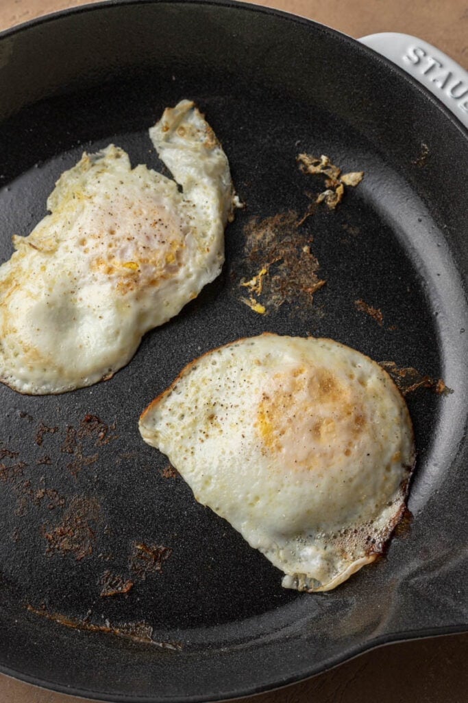 Two fried eggs cooking in a black nonstick skillet, with slightly crispy edges and golden spots.