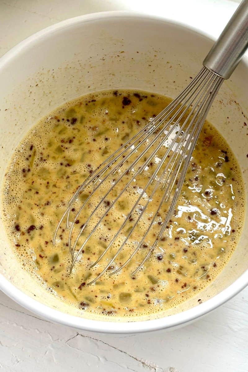 A metal whisk in a bowl of seasoned egg mixture with visible spices and chopped vegetables.