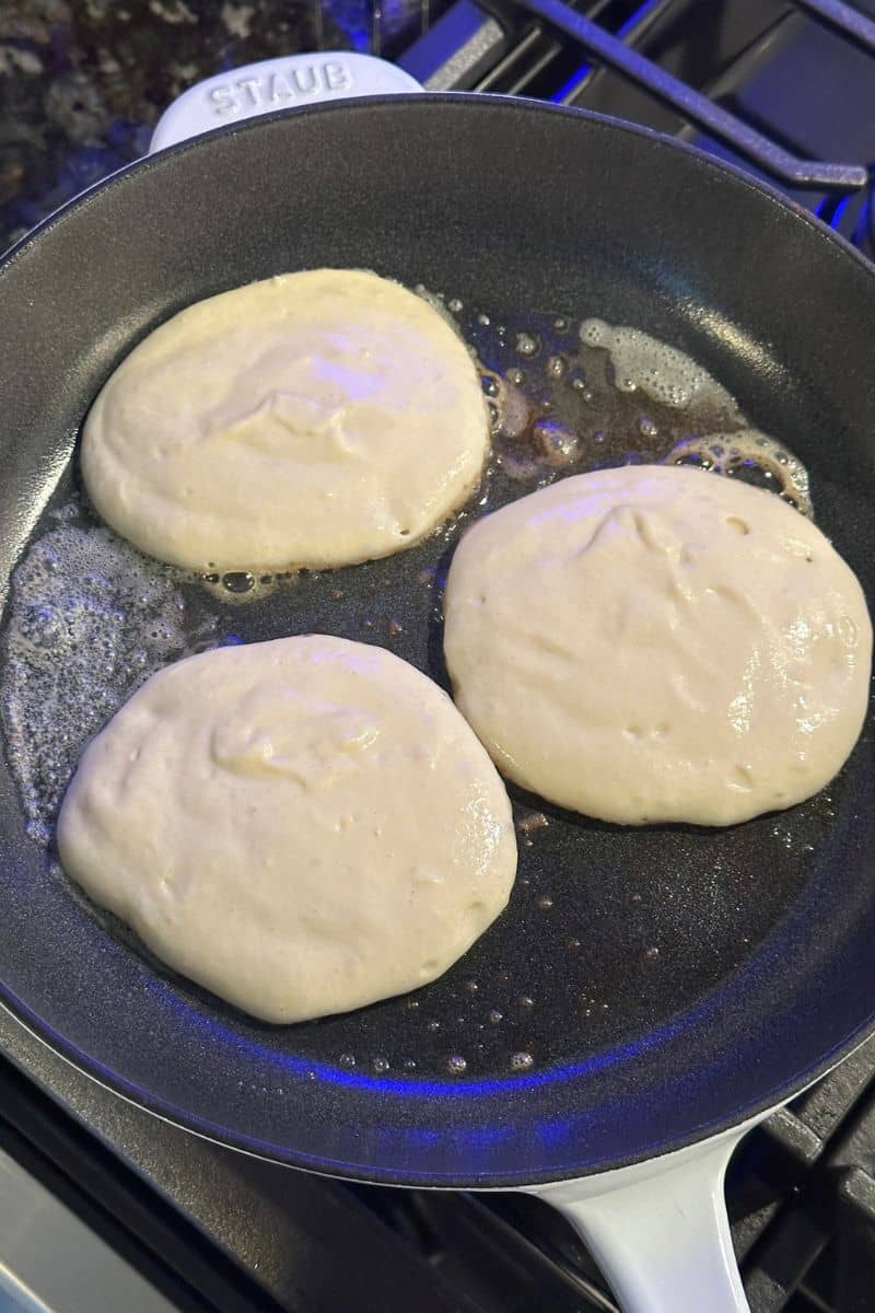 Three pancakes cooking in a frying pan on a stovetop with bubbling butter around them.