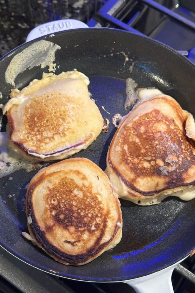 Three golden-brown pancakes cooking in a black pan on a stove, with some batter spilled on the sides.