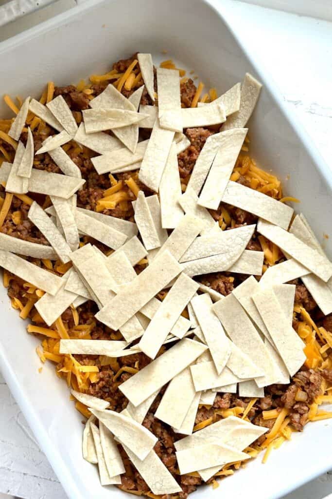 Rectangular baking dish with ground meat, shredded cheese, and uncooked tortilla strips layered inside.