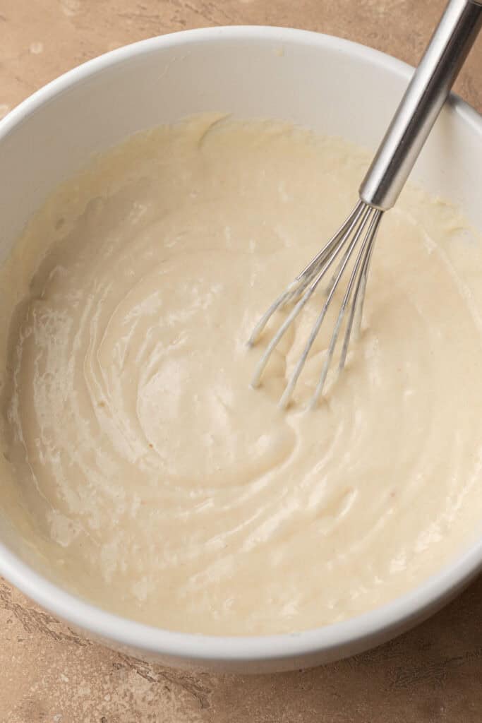A metal whisk in a white bowl of smooth, creamy batter on a beige surface.