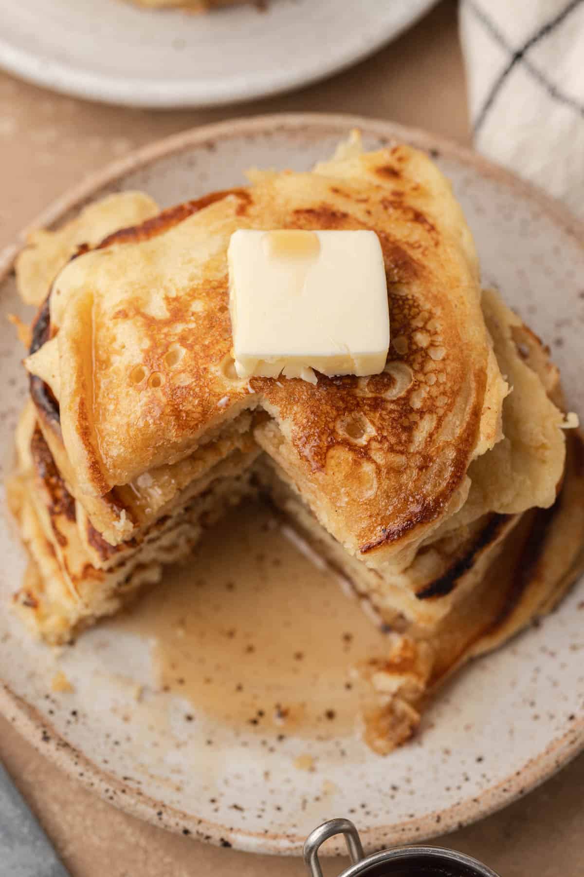 Stack of pancakes with a pat of butter on top, syrup drizzled, and a wedge cut out on a speckled plate.