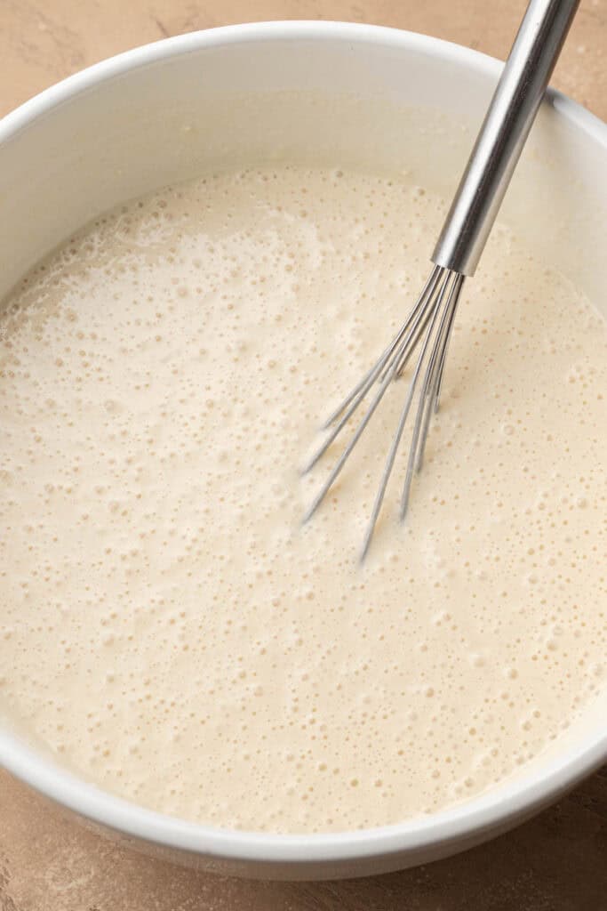 A metal whisk in a white bowl filled with bubbly, smooth pancake batter.