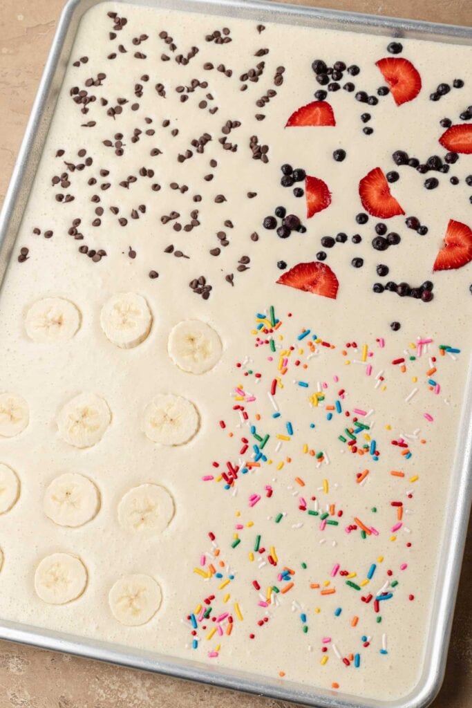 Sheet pan pancake batter topped with chocolate chips, berries, banana slices, and rainbow sprinkles.