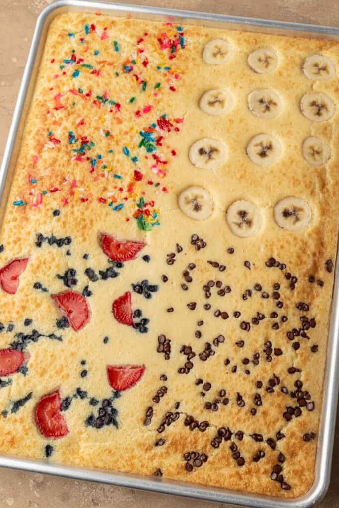 Sheet pan pancake topped with sections of sprinkles, banana slices, strawberries, blueberries, and chocolate chips.