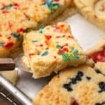 A spatula lifts a square of confetti cake from a tray of assorted fruit-topped cake squares.