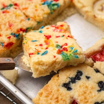A spatula lifts a square of confetti cake from a tray of assorted fruit-topped cake squares.