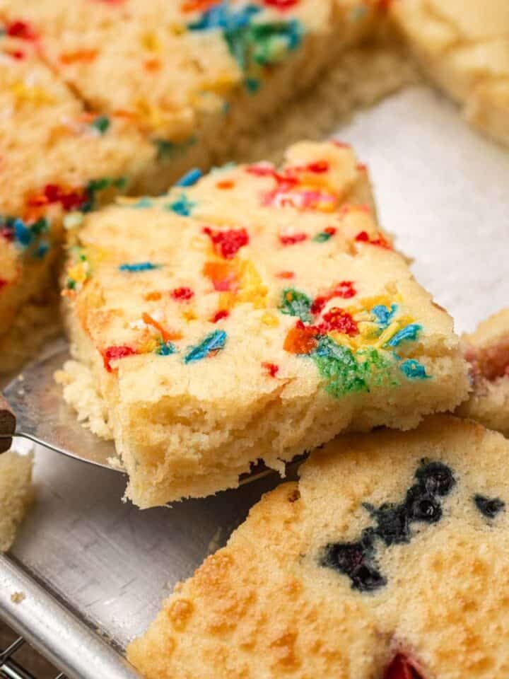A spatula lifts a square of confetti cake from a tray of assorted fruit-topped cake squares.